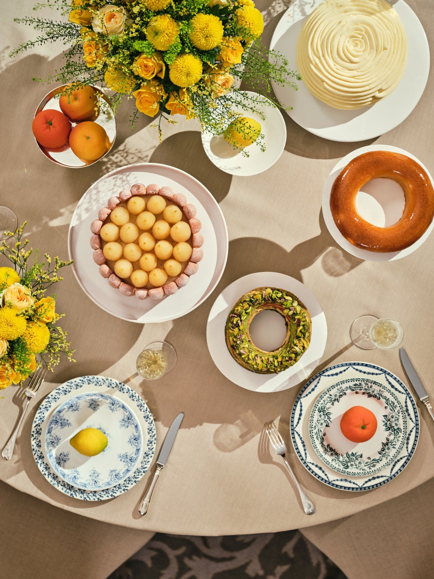 Uma mesa redonda com pratos elegantes, sobremesas e frutas. Flores amarelas estão dispostas num bouquet. O ambiente é acolhedor e convidativo, evocando um clima festivo.