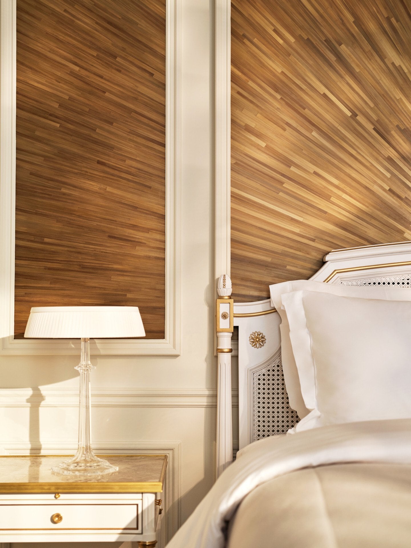 Canto do lado direito da cama, ao lado de uma mesa de cabeceira e de um candeeiro em cima, e a parede feita com marchetaria de palha, em Le Meurice, Paris