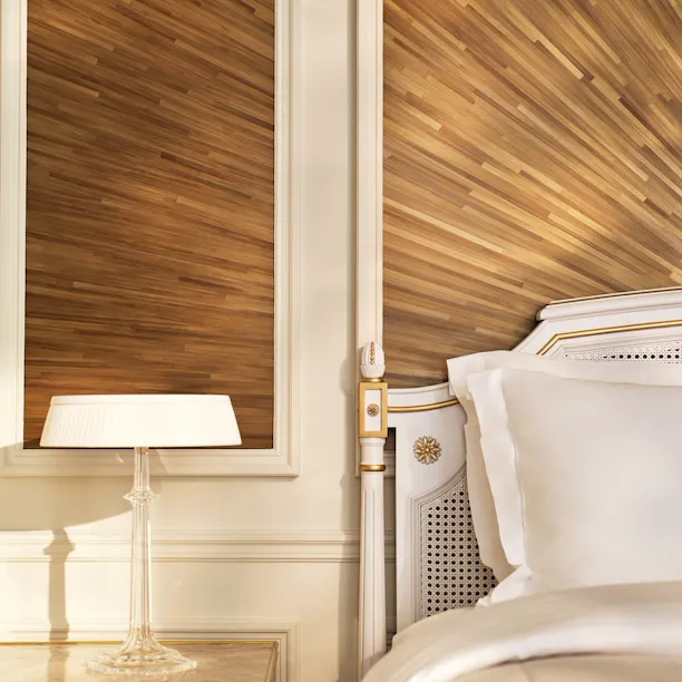 Canto do lado direito da cama, ao lado de uma mesa de cabeceira e de um candeeiro em cima, e a parede feita com marchetaria de palha, em Le Meurice, Paris