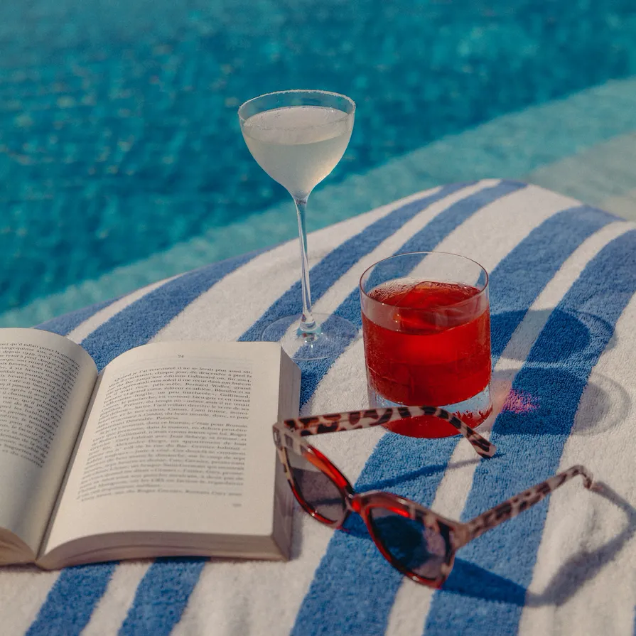 A close up to a book, a pair of sunglasses and two cocktails on High Society's sunbed.