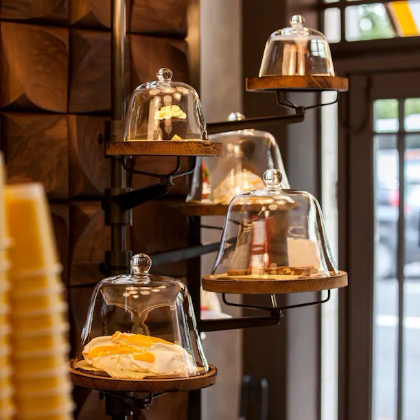 Display of freshly prepared pastries at Parcafé at The Dorchester, showcasing cakes and sweet treats arranged on tiered wooden shelves beneath glass cloches, illuminated by warm lighting and set against rich wood panelling.