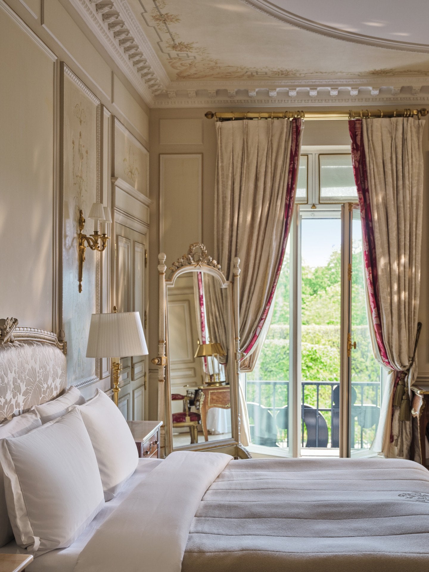 Vista ampla do lado direito da cama com o espelho ao fundo e as janelas e cortinas abertas, no Le Meurice, Paris
