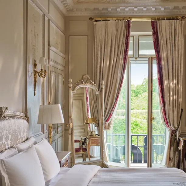 Vista ampla do lado direito da cama com o espelho ao fundo e as janelas e cortinas abertas, no Le Meurice, Paris