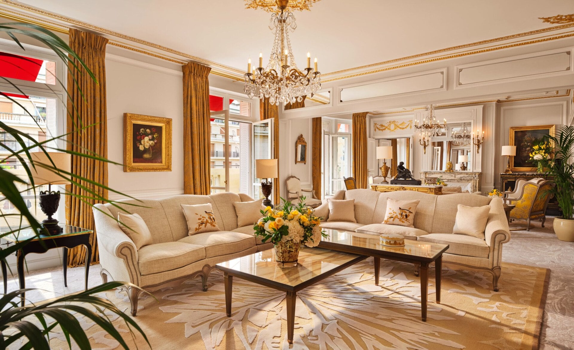 Sala de estar luxuosa com sofás elegantes em tom creme, mesa de centro em vidro e detalhes dourados. Um lustre e janelas amplas criam um ambiente luminoso e opulento.