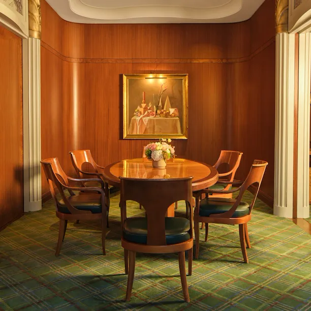 A round wooden table with six chairs in a wood-paneled room with framed art and floral centerpiece