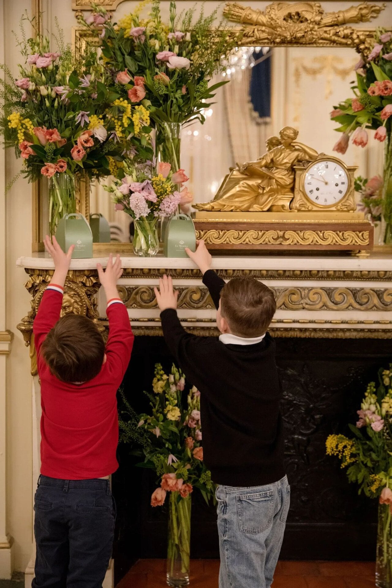 Two young children, one in a red sweater and the other in a black one, stand in front of an ornate fireplace, reaching up towards the mantel. The mantel is decorated with large flower arrangement and a golden clock with a reclining figure. At Le Meurice, Paris.