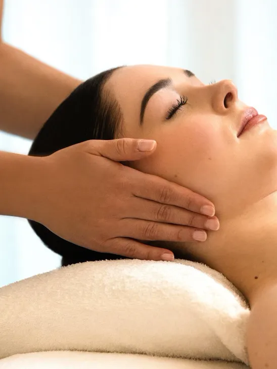 We can see a woman lying on a massage table, resting her head on a white pillow. We can see her from the side. The masseuse's hands are placed on the client's ears. The client's eyes are closed at the spa Valmont at Le Meurice, Paris.