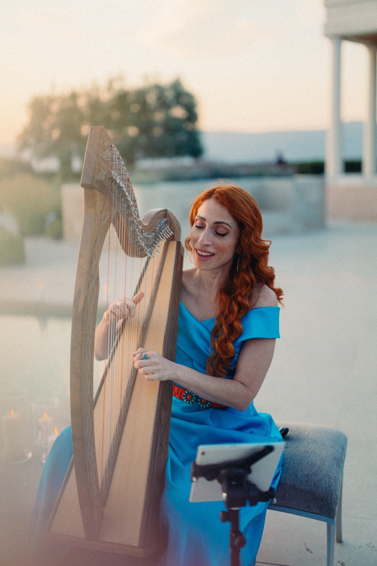Une femme aux longs cheveux roux vêtue d'une robe bleue joue de la harpe en plein air au coucher du soleil. Elle sourit chaleureusement, créant une atmosphère sereine et joyeuse.