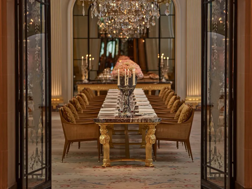 Jean Imbert au Plaza Athénée's central marble table, drawned up with pink flowers and chandeliers. Crystal chandeliers hanging above the table. Wrought iron and glass door at the forefront. Hotel Plaza Athénée, Paris