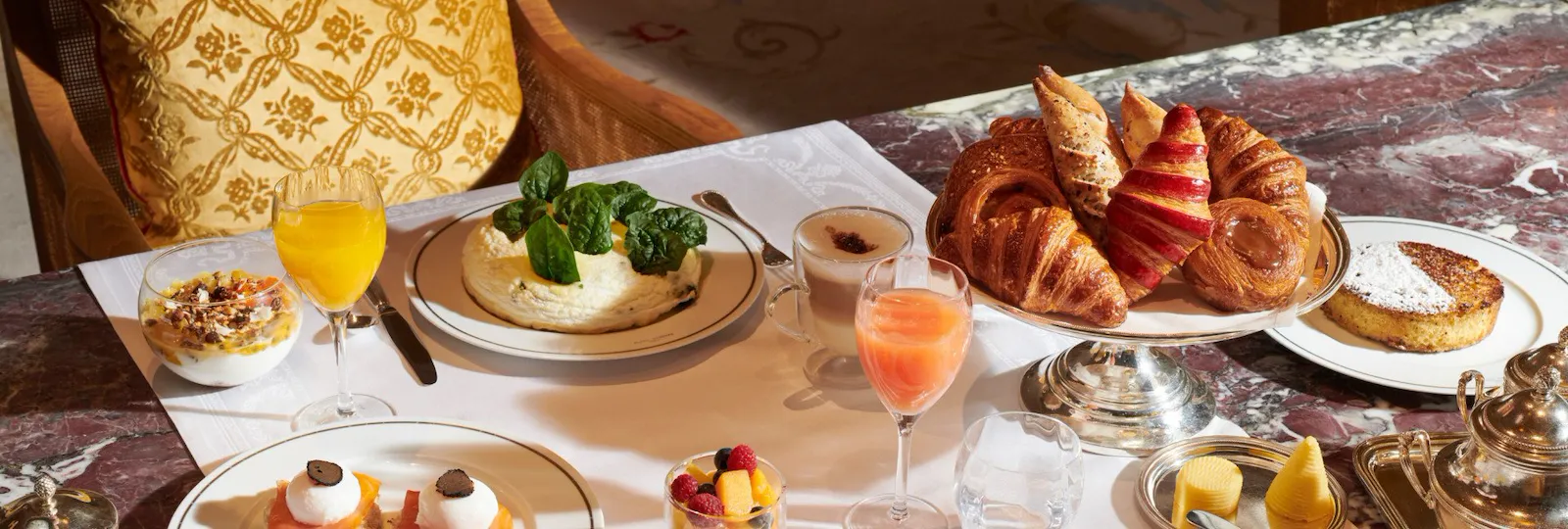 Imagem do brunch no Jean Imbert au Plaza Athénée. Podemos ver uma salada de frutas, bolos, café, ovos, crepes, abacate e sucos