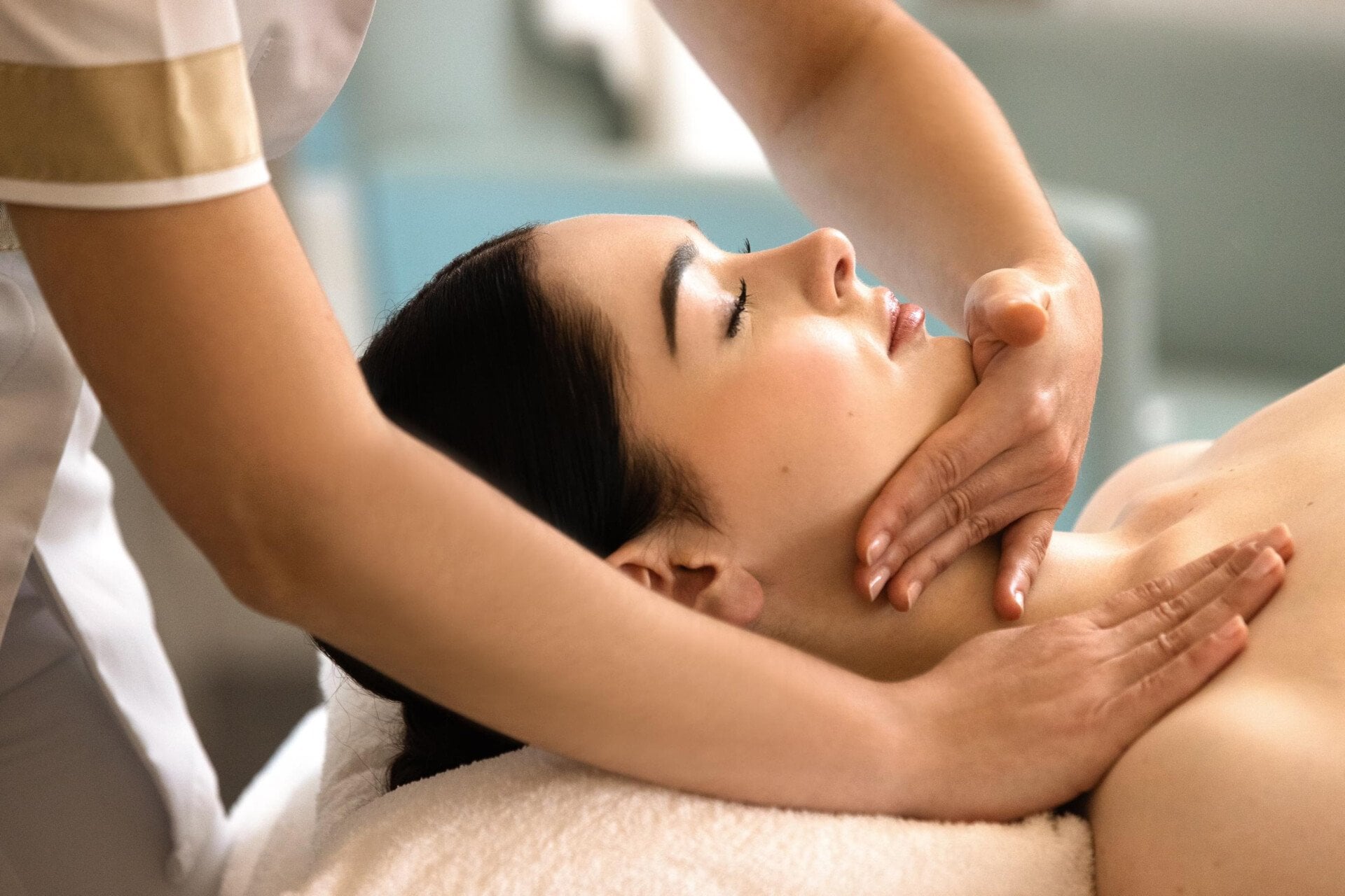 In the photo you can see a customer's head resting on a white cushion. The client is lying on the massage table.  Her eyes are closed. We can see both hands and one sleeve of the masseuse's uniform. One of these hands is on the client's chin and neck. We can see the delicacy of the masseuse's hands. One of these hands is located on the client's upper chest at Le Meurice, Paris.