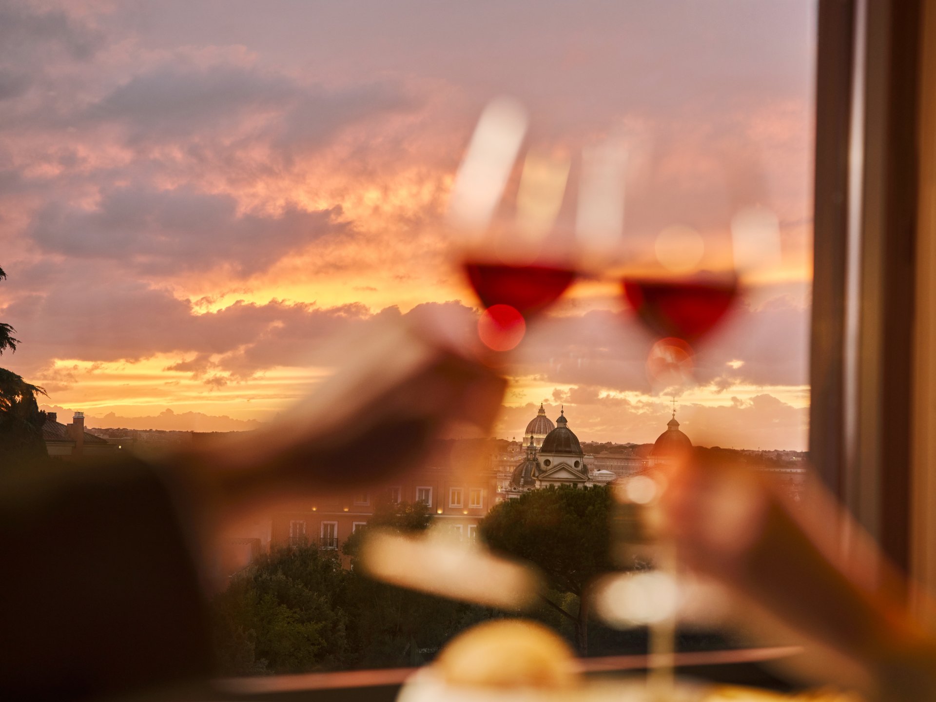 Due bicchieri di vino rossi avvicinati per un brindisi con lo sfondo di un tramonto su Roma