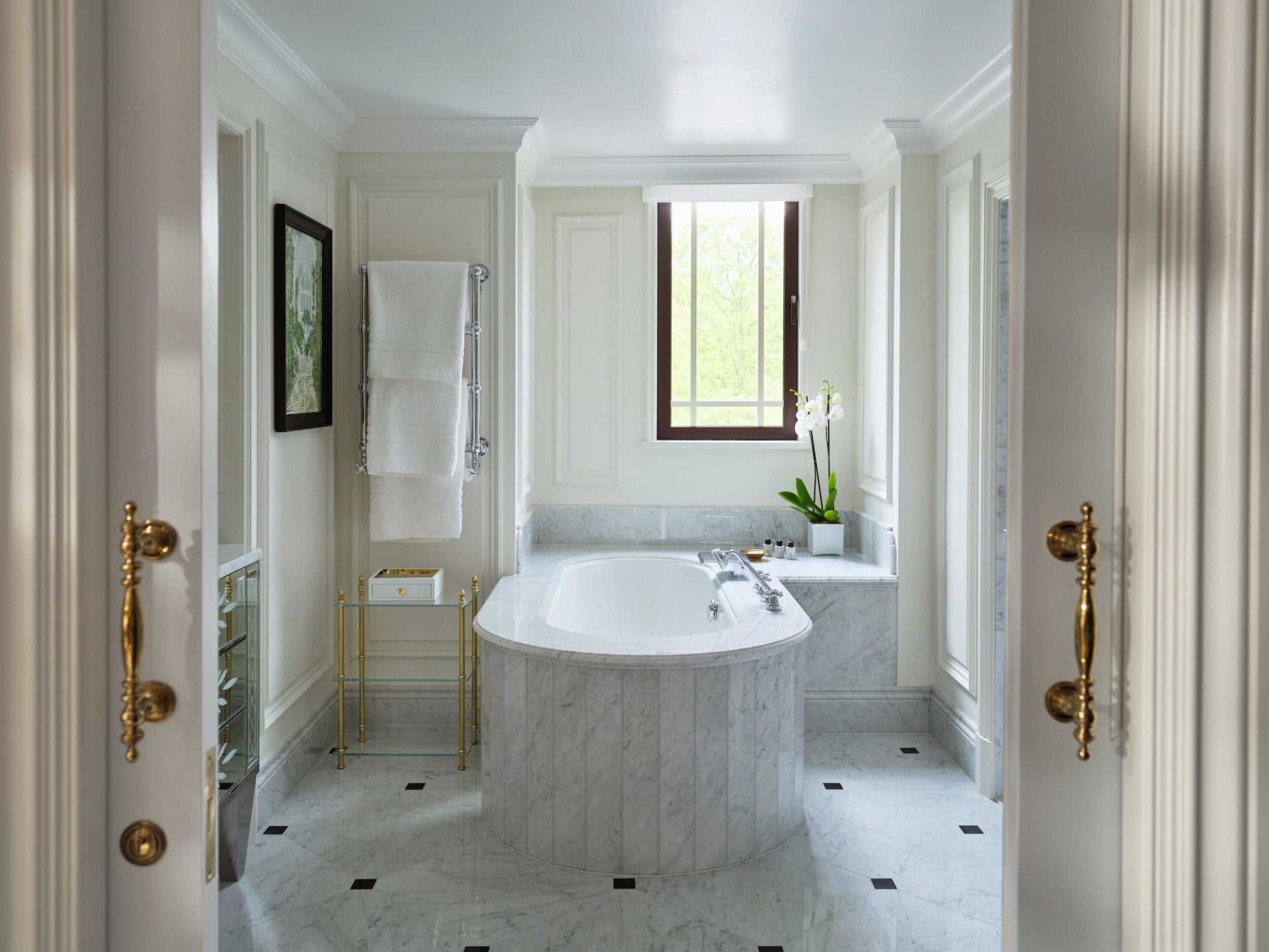 Bathroom in The Hyde Park Suite with a freestanding marble bath, white marble flooring, panelled walls, a towel stand and a large window at the far end of the room.