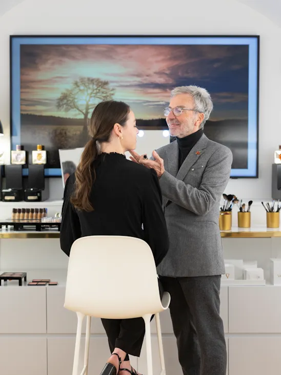 Un homme en costume gris maquille une femme assise sur une chaise blanche. Ils se trouvent dans un studio de maquillage moderne, avec des produits de beauté et une grande photo de paysage en arrière-plan. L'atmosphère est professionnelle et concentrée.