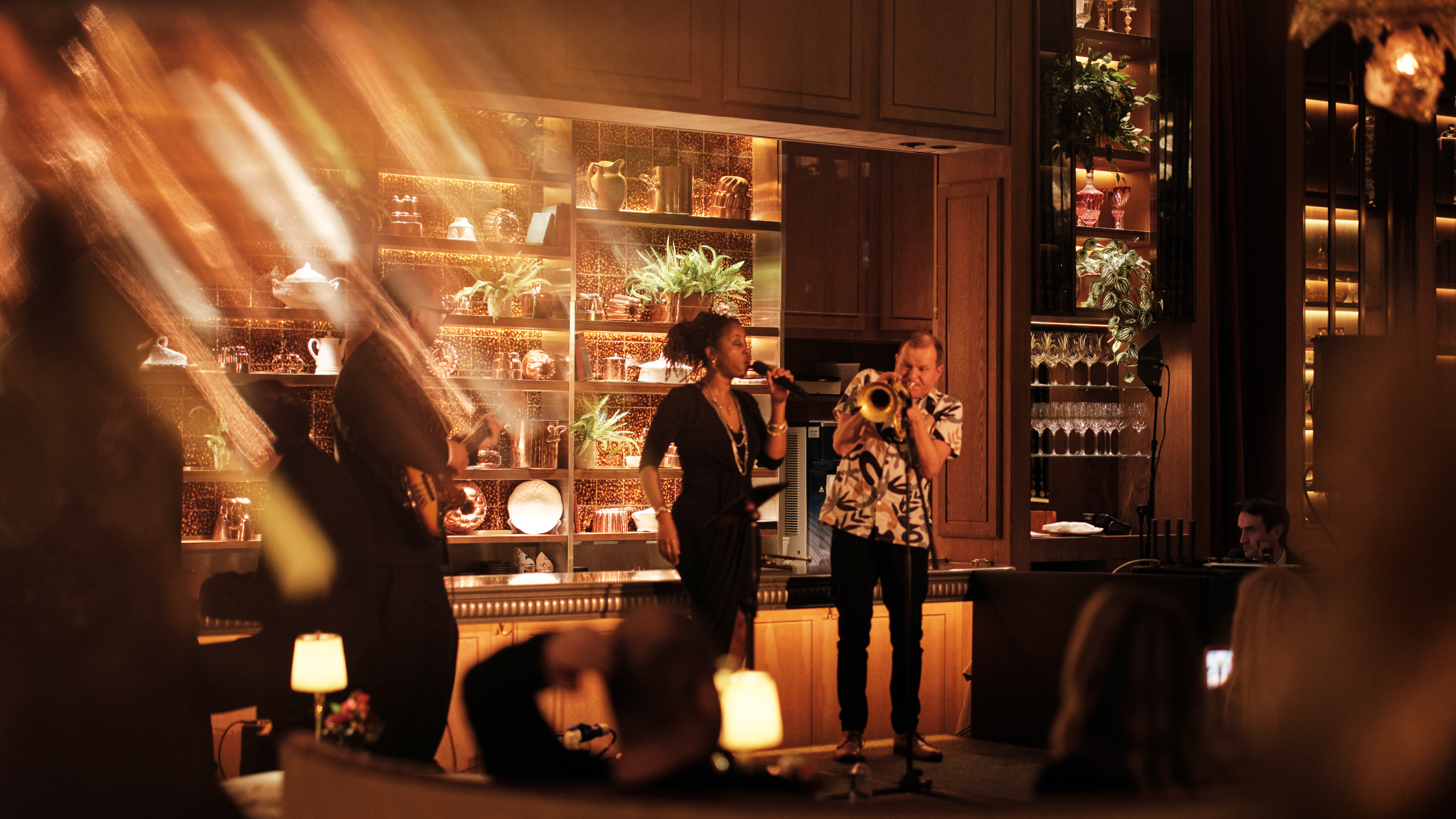 Live music at Ronnie Scott’s at The Dorchester, showcasing musicians performing on stage in front of a mirrored back bar, with warm lighting and guests seated throughout the intimate venue.