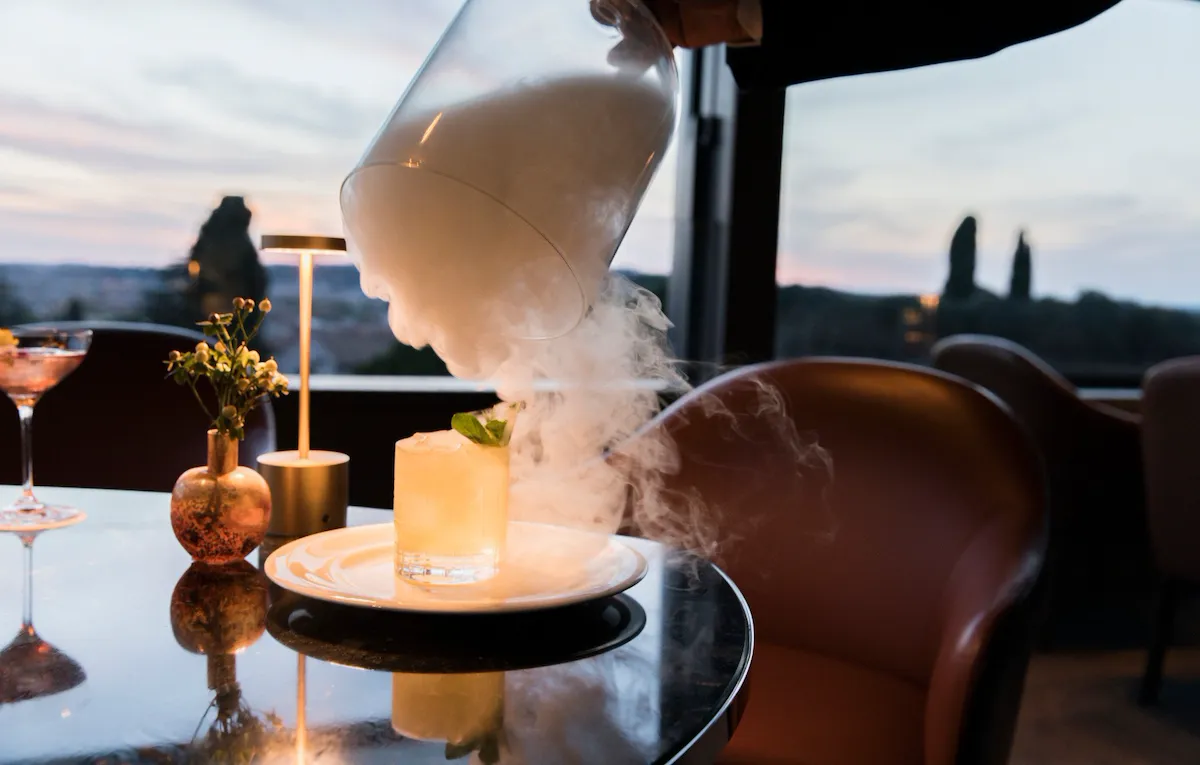 Cocktail appoggiato su di un tavolo all'aperto a Il Giardino bar. La mano di un barman solleva una campana di vetro da cui esce fumo bianco