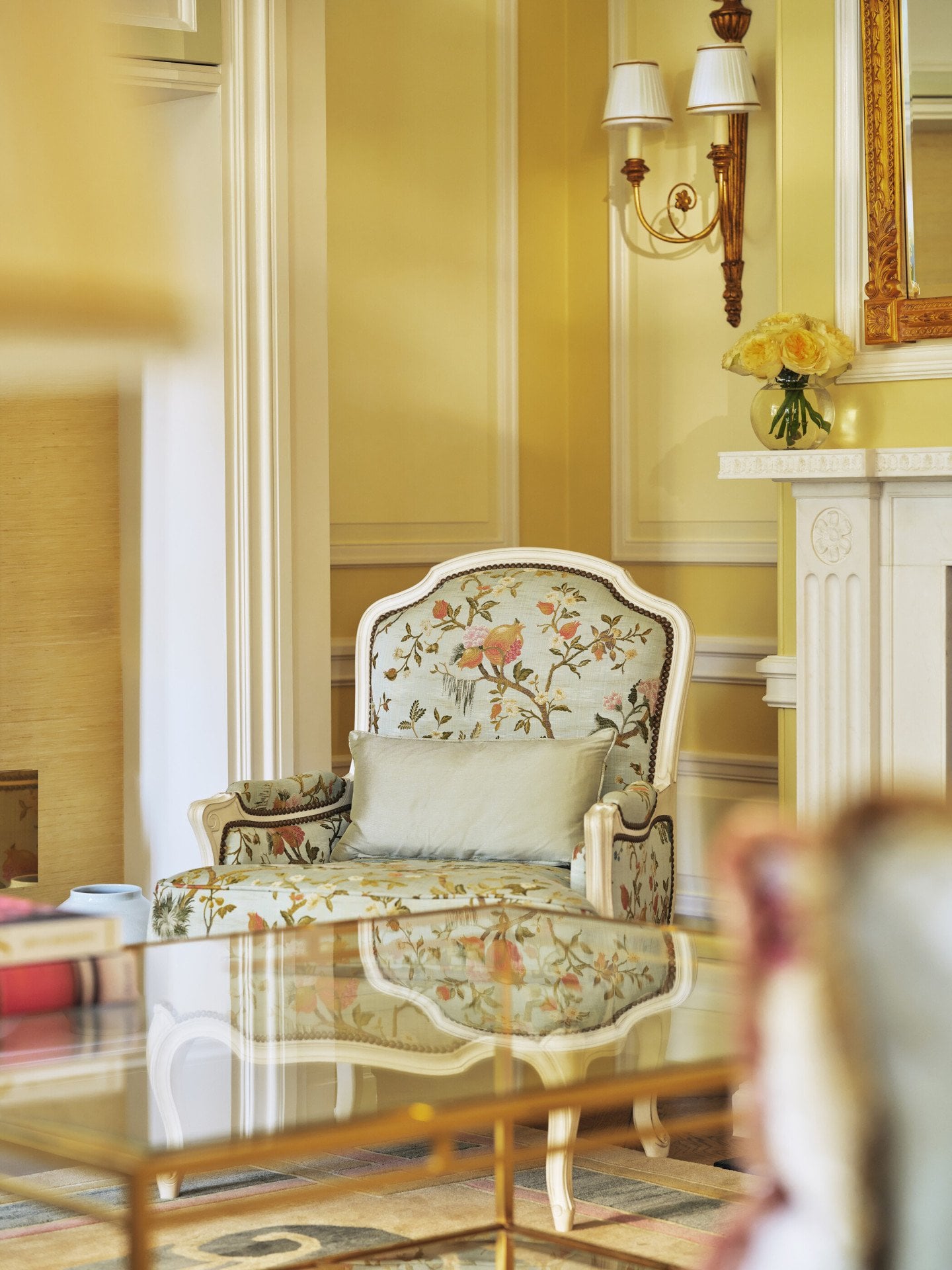 The Mayfair Suite at The Dorchester, showcasing a floral-patterned armchair, a glass-topped coffee table, soft yellow walls, a decorative fireplace and fresh cream roses displayed beneath a gilded mirror.