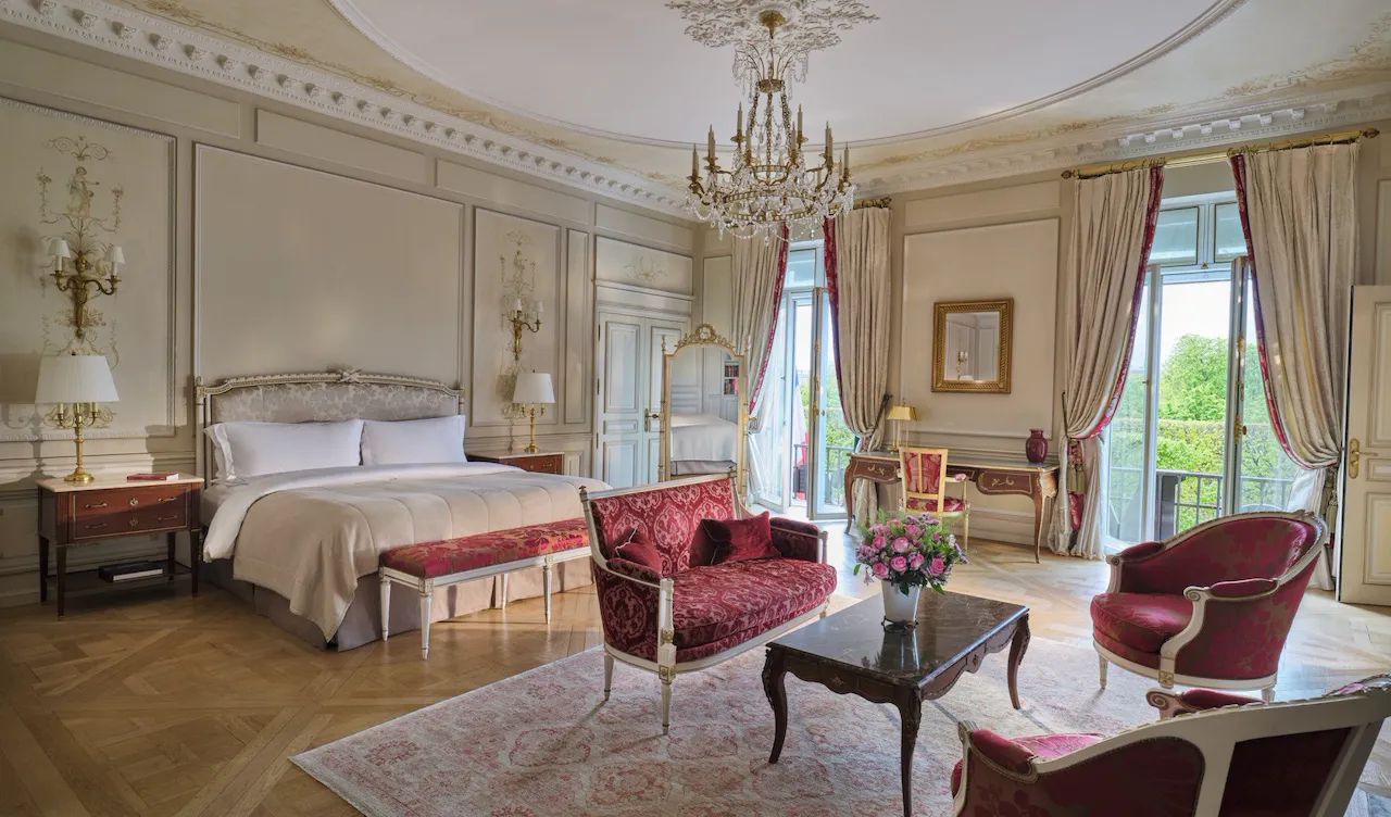 Vista ampla da Suíte Presidencial Dali com em primeiro plano os móveis de uma sala de estar, como sofá, carpete e cadeiras, e ao fundo a cama e o espelho, no Meurice, Paris