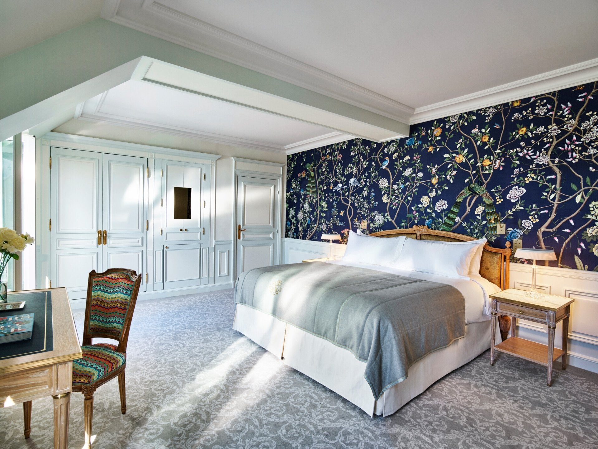 Um quarto luxuoso com uma cama grande com edredão cinzento e almofadas brancas. O papel de parede floral azul confere elegância. Os guarda-roupas brancos e uma cadeira estampada realçam a sensação de luminosidade e amplitude do quarto. A luz suave entra, criando uma atmosfera serena.