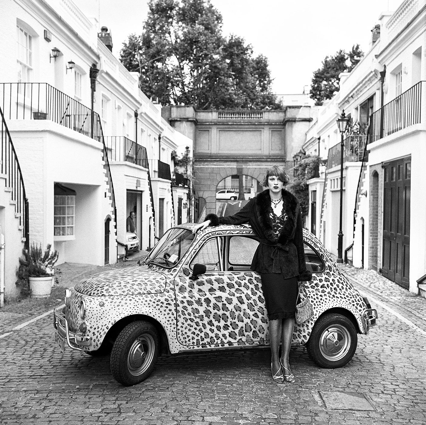 Person stands beside a vintage car on a quiet city street in black and white.