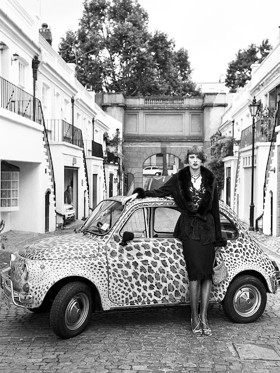 Person stands beside a vintage car on a quiet city street in black and white.