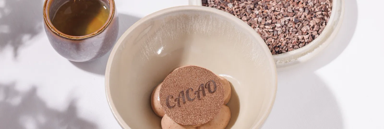 Mousse al cacao in una ciotola su di un tavolo accanto ad una tazzina con una bevanda al cacao e un piatto con un cioccolatino a forma di fava di cacao