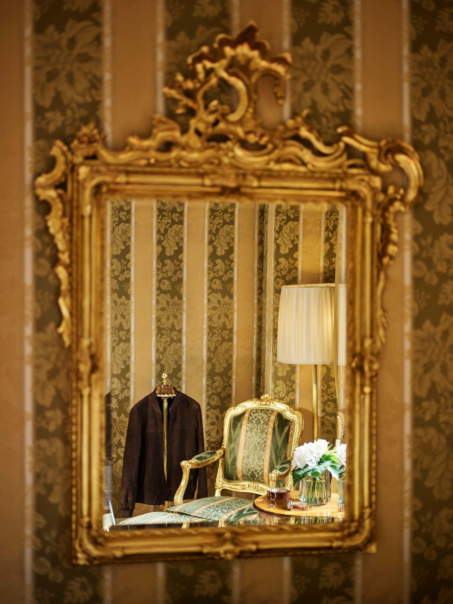 Riflesso dello specchio della camera da letto della Royal Suite che mostra la particolare carta da parati dai colori verde e oro, un elegante poltrona a fianco di un tavolo con un vaso di fiori