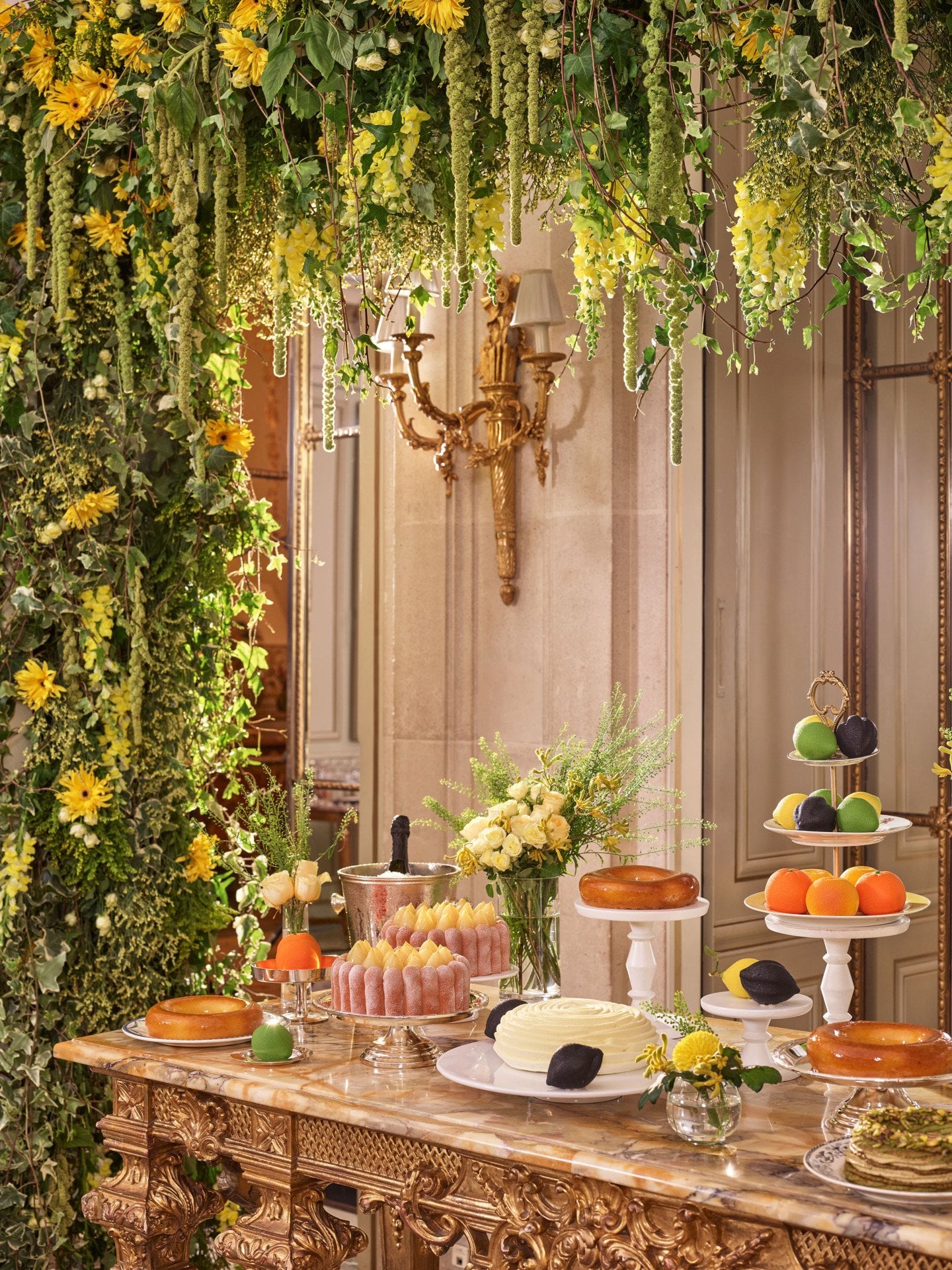 Mesa de sobremesas luxuosa com bolos e doces em móveis dourados ornamentados. No teto, vegetação exuberante e flores amarelas criam um ambiente vibrante e elegante.