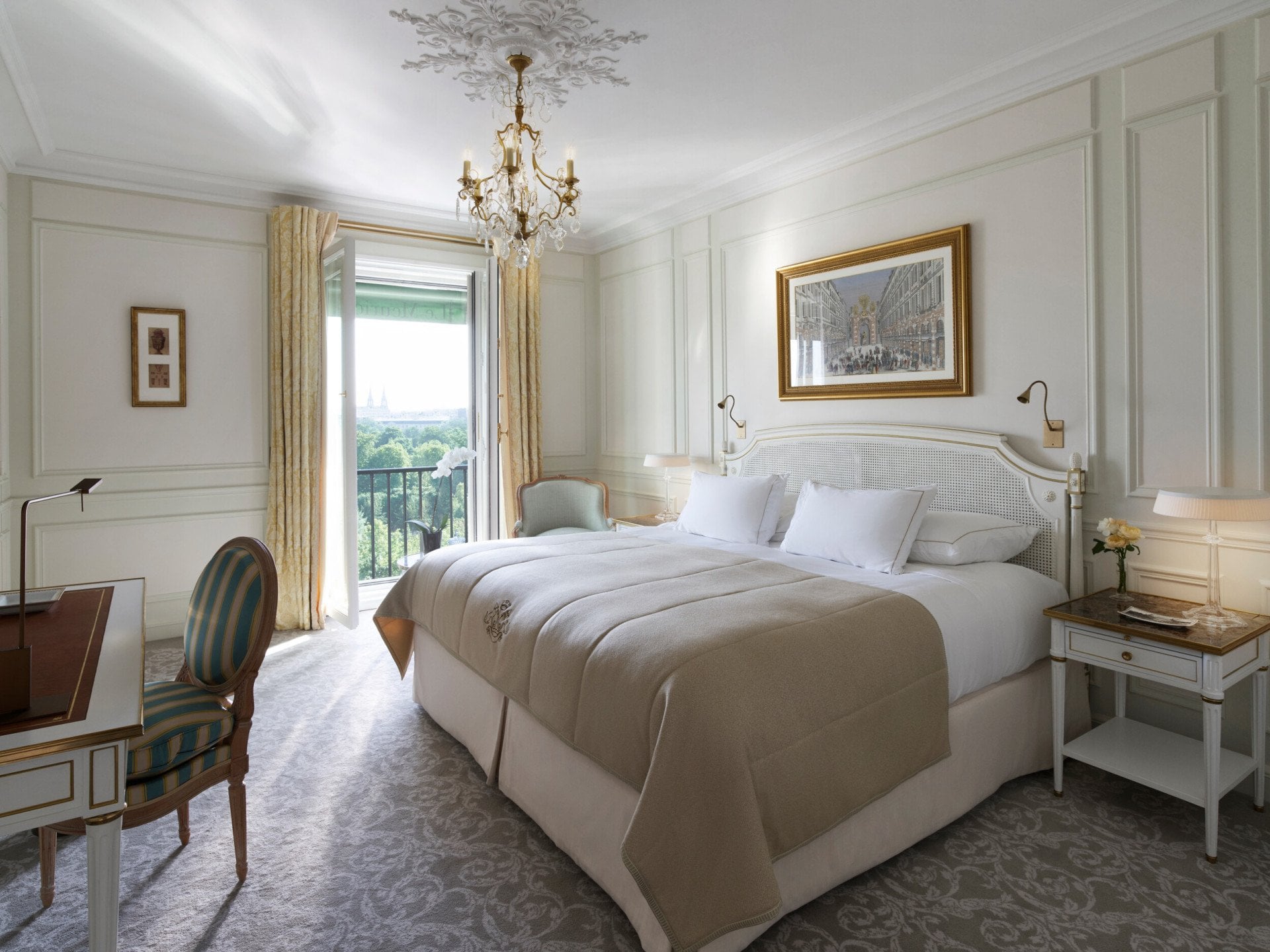 Quarto luxuoso com cama grande, roupa de cama bege e almofadas brancas. Um lustre pendurado no teto. As portas da varanda abertas revelam uma vista exuberante.