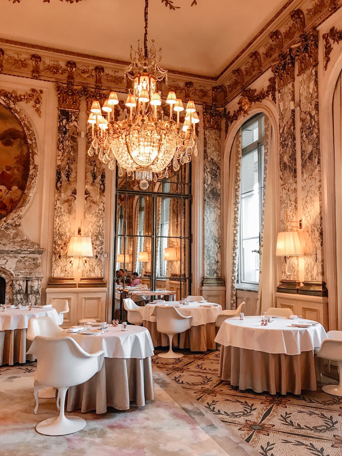 Restaurant room with a focus on a large chandelier lighting the gold ornaments on the walls. At Le Meurice, Paris.