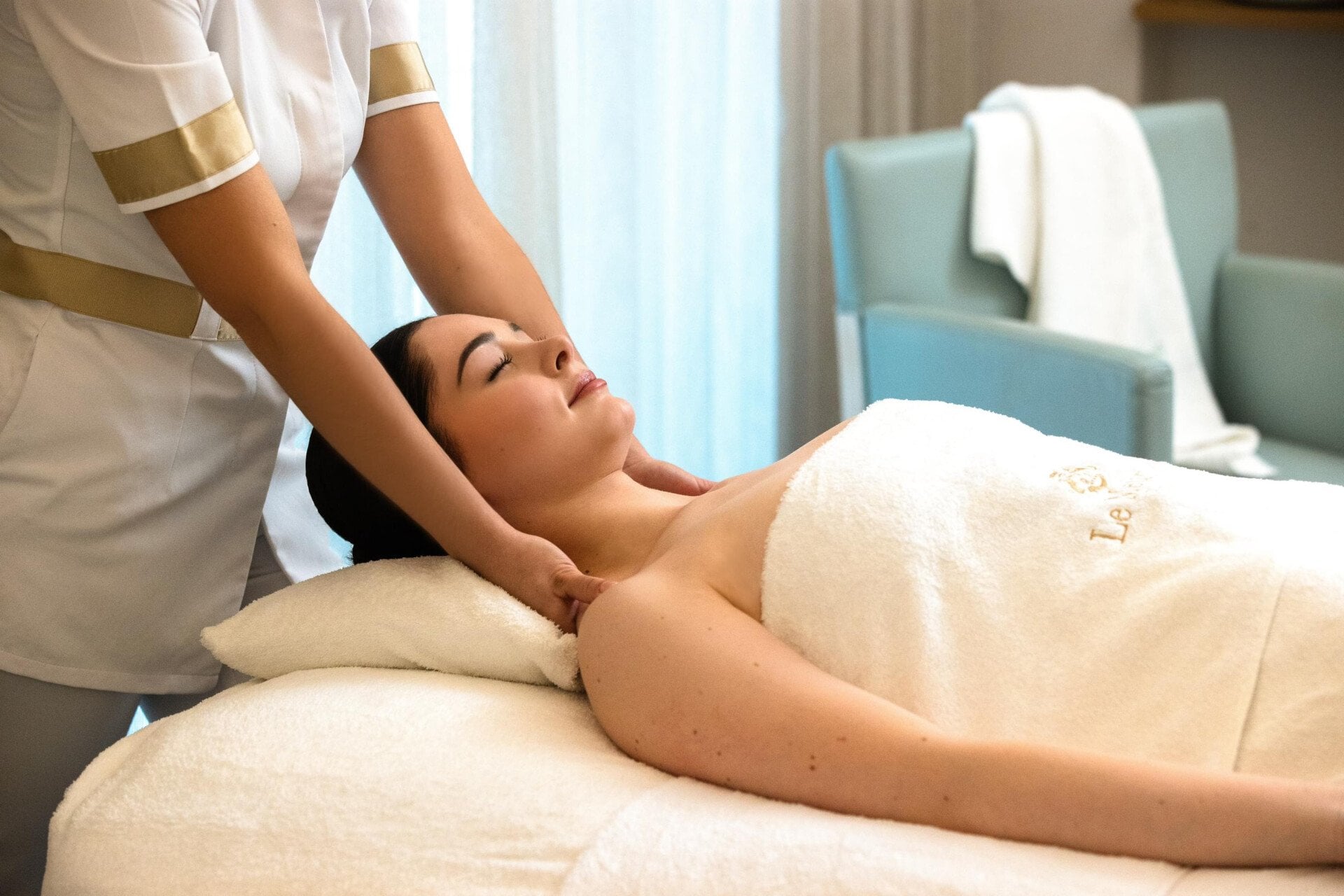 On this picture, we can see the client lies on the massage table resting her head on a white pillow.  We observe the client from the side. Her upper chest is naked, the rest of her body hidden by a Le Meurice towel. On the left, we can see the masseuse's uniform. The masseuse has her arms on the client's shoulders. In the background, we can see a blue armchair with a white towel on top at Le Meurice, Paris.