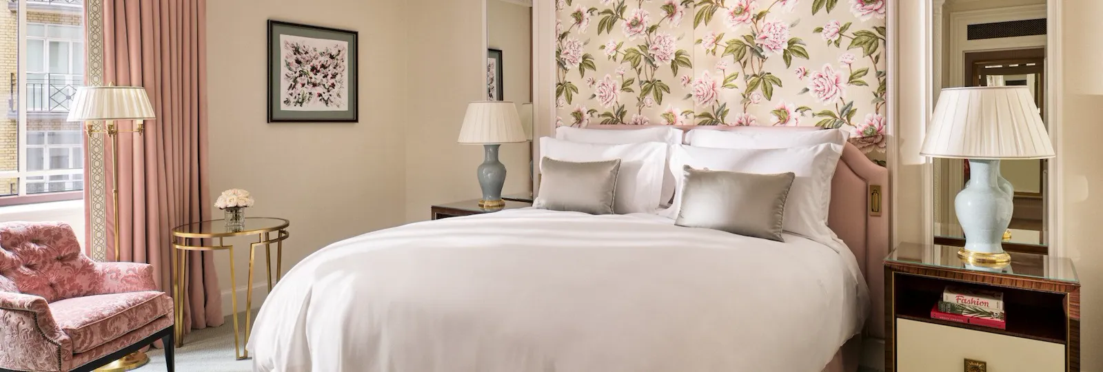 The Superior Room at The Dorchester, showcasing a king-sized bed framed by botanical floral wall coverings, blush-pink upholstery, classic bedside tables and soft natural light from the window.