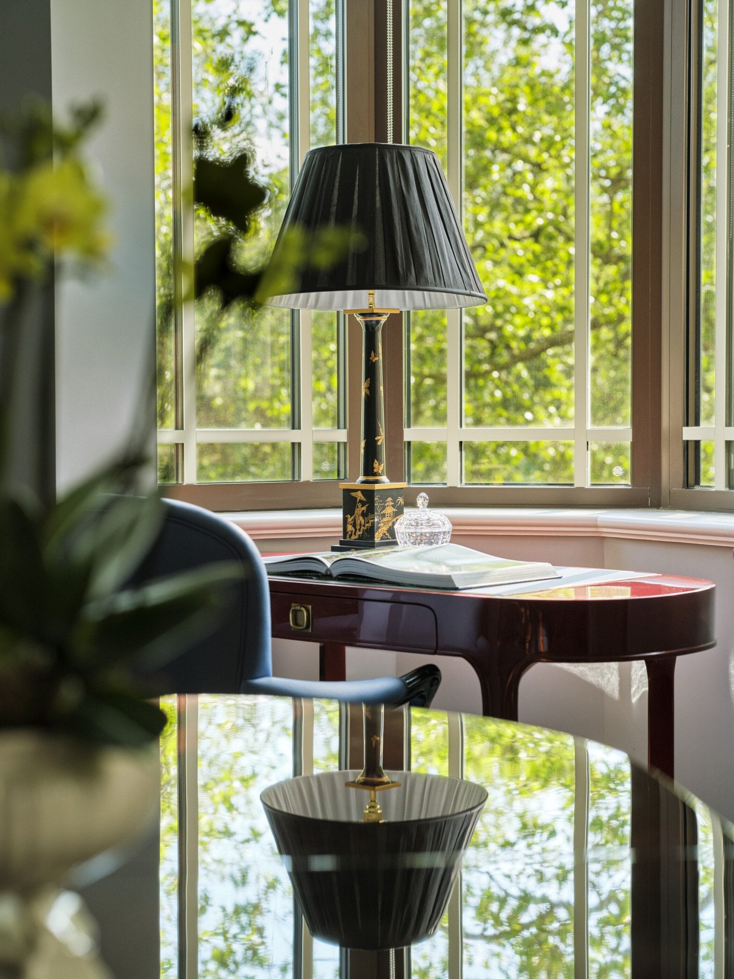 The Regal Suite at The Dorchester, featuring an elegant wooden writing desk, a black table lamp and expansive bay windows that fill the room with natural light and views of Hyde Park.