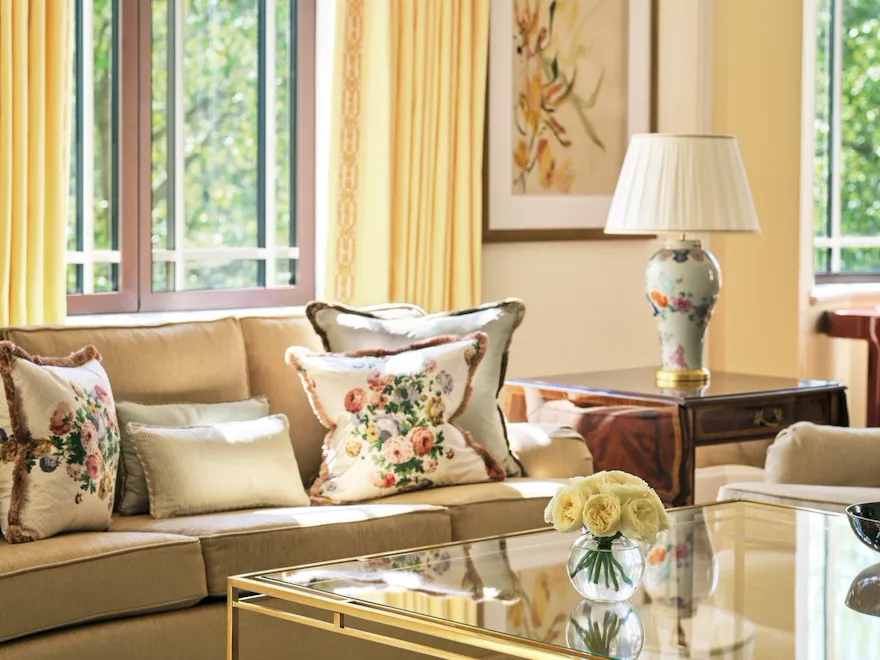 The Dorchester Suite living room at The Dorchester, showcasing comfortable lounge seating, floral accent cushions, a glass coffee table, soft yellow drapery and large windows framing views of Hyde Park and Park Lane.