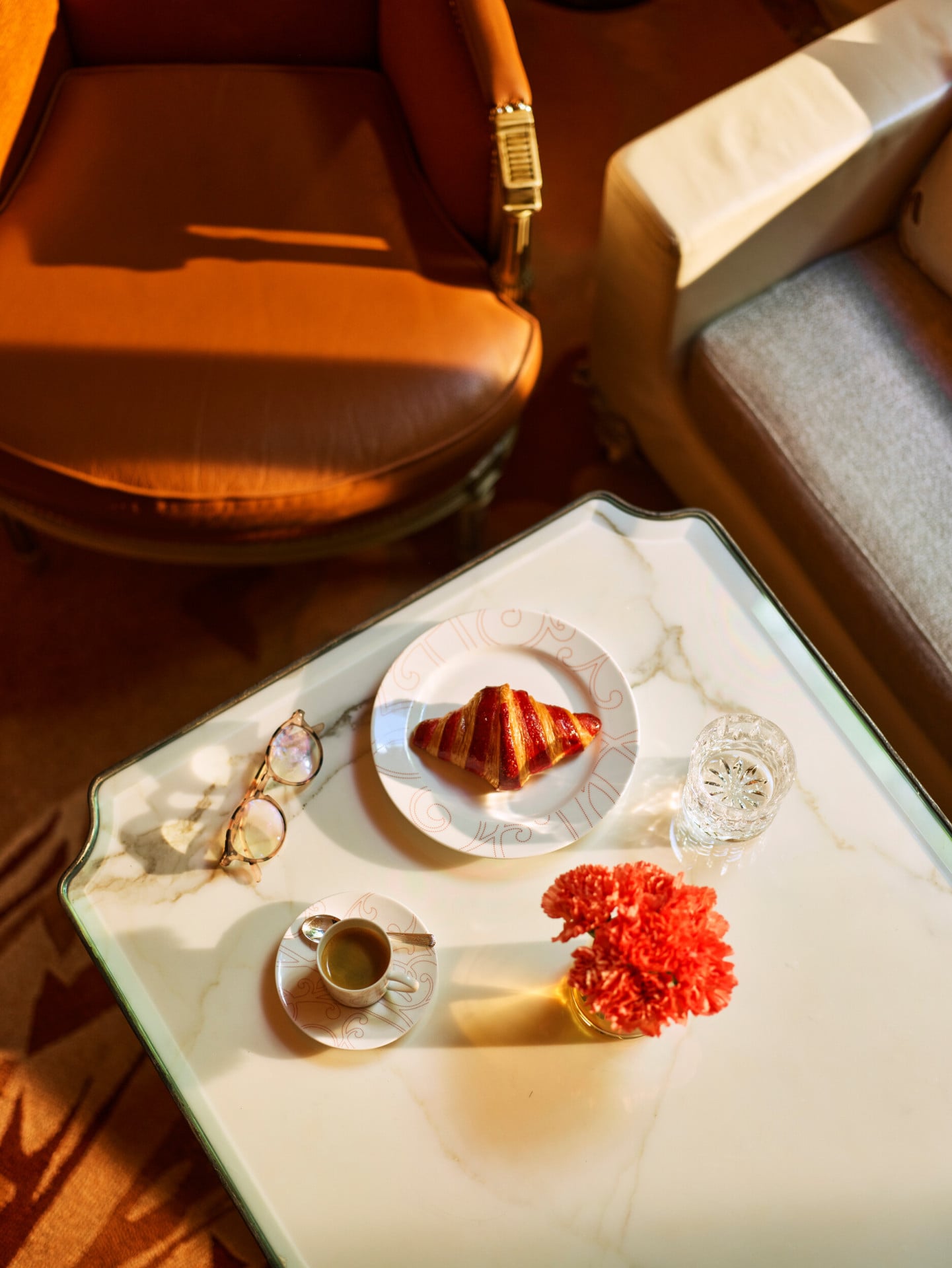 Mesa de jantar em mármore branco na galeria vista de cima, com óculos de leitura, uma xícara de café, um croissant num prato e um gerânio vermelho, no Hotel Plaza Athénée, Paris