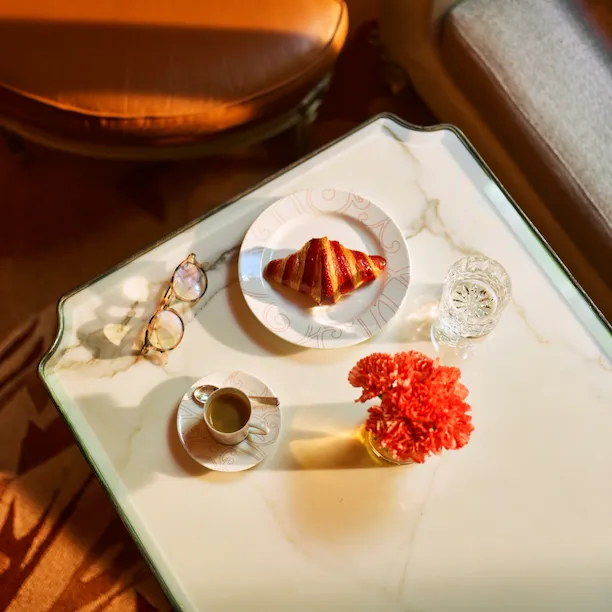 Mesa de jantar em mármore branco na galeria vista de cima, com óculos de leitura, uma xícara de café, um croissant num prato e um gerânio vermelho, no Hotel Plaza Athénée, Paris