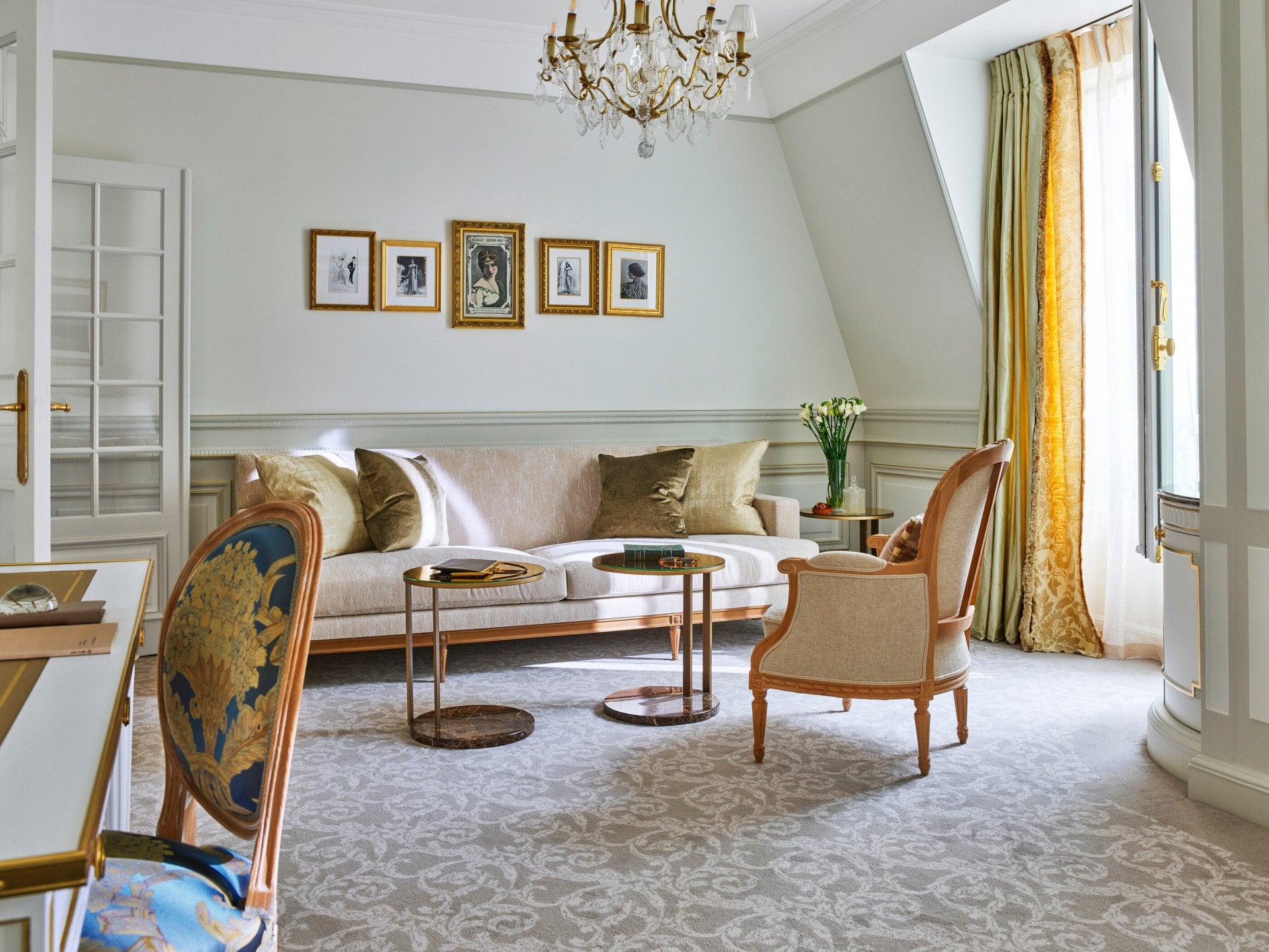 Sala de estar elegante com sofá bege, almofadas decorativas e duas mesas redondas. Um lustre dourado pendurado no teto. Quadros decoram as paredes.