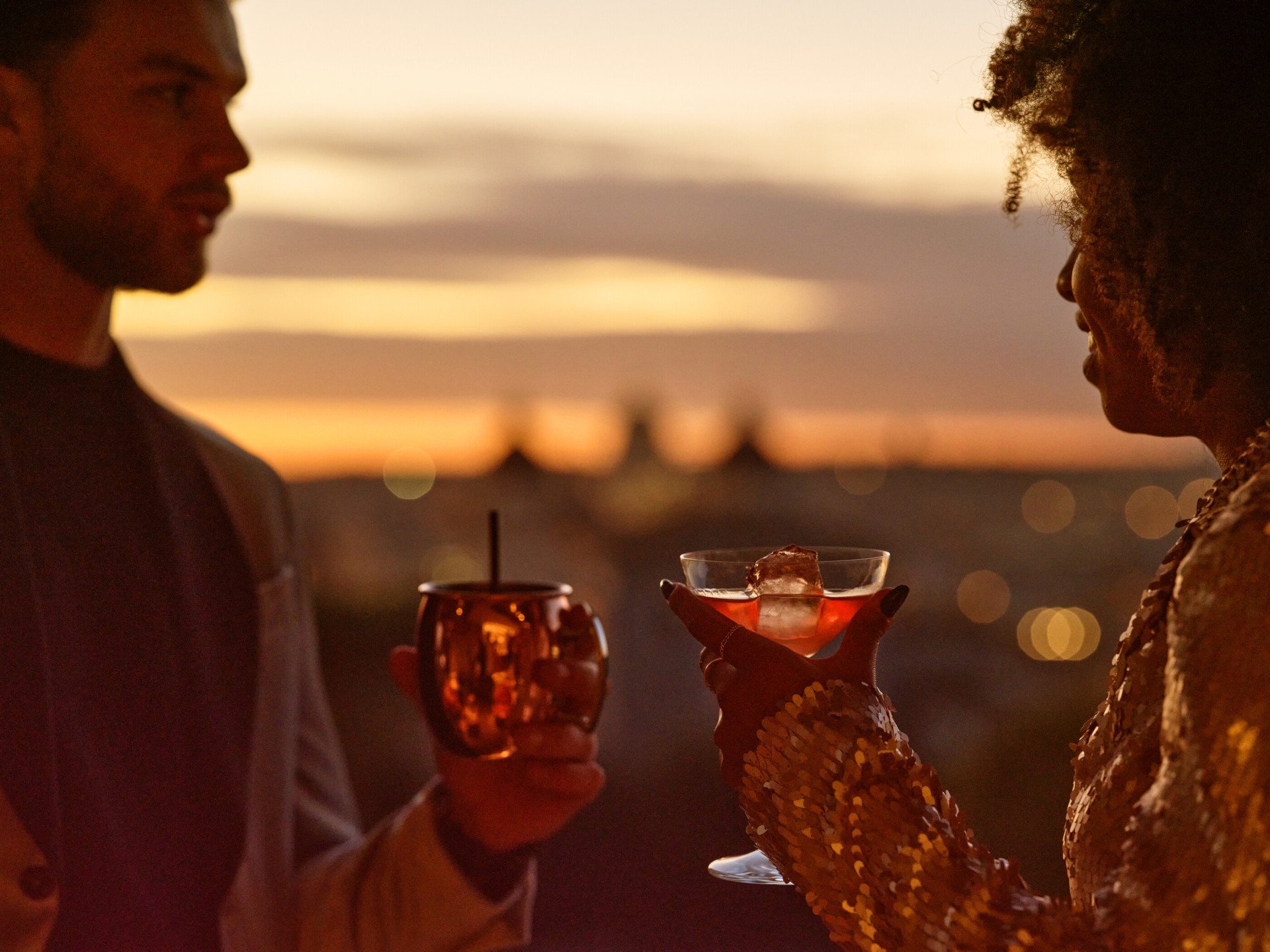 Un giovane uomo e una giovane donna con due drink che parlano con lo sfondo di un tramonto su Roma
