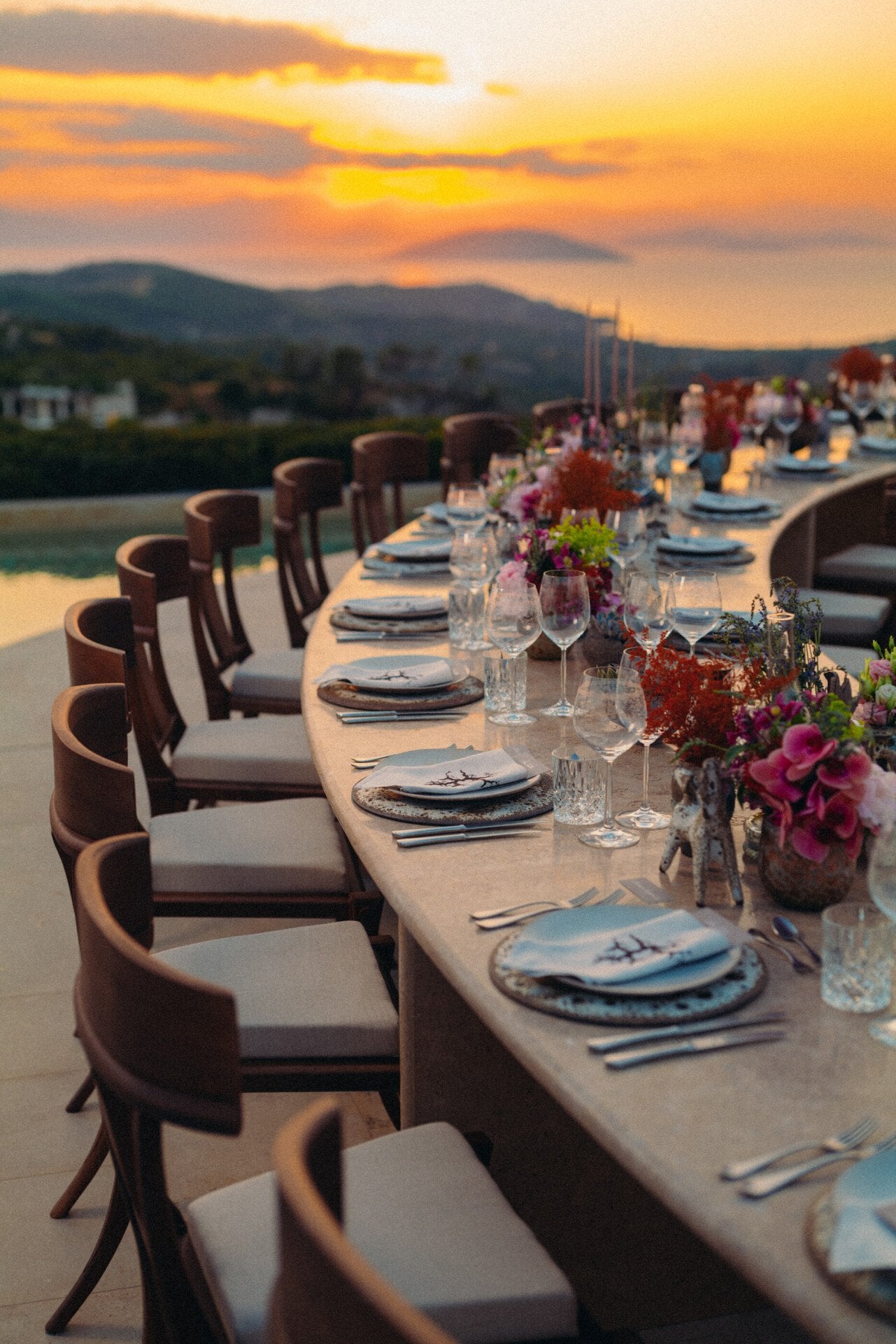 Élégant décor pour un dîner en plein air avec une longue table, ornée de fleurs colorées et d'une vaisselle raffinée, avec en toile de fond un coucher de soleil éclatant sur les collines vallonnées.