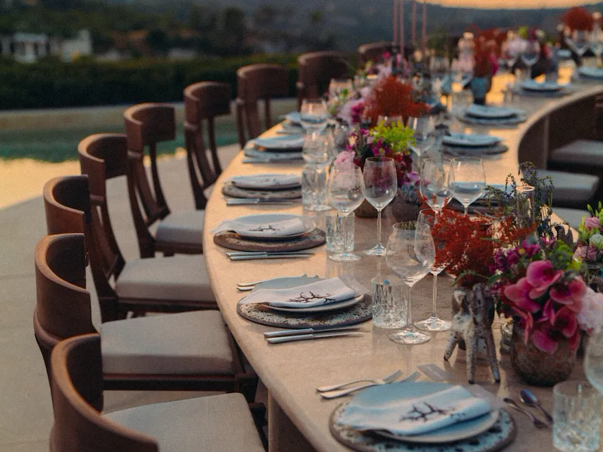 Élégant décor pour un dîner en plein air avec une longue table, ornée de fleurs colorées et d'une vaisselle raffinée, avec en toile de fond un coucher de soleil éclatant sur les collines vallonnées.