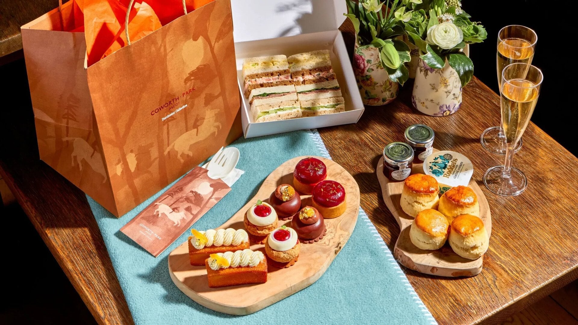 Takeaway meadow afternoon tea with finger sandwiches in box, summer pastries, scones, cream and jam and champagne layed out on picnic blanket with takeaway bag for the Royal Windsor Flower Show