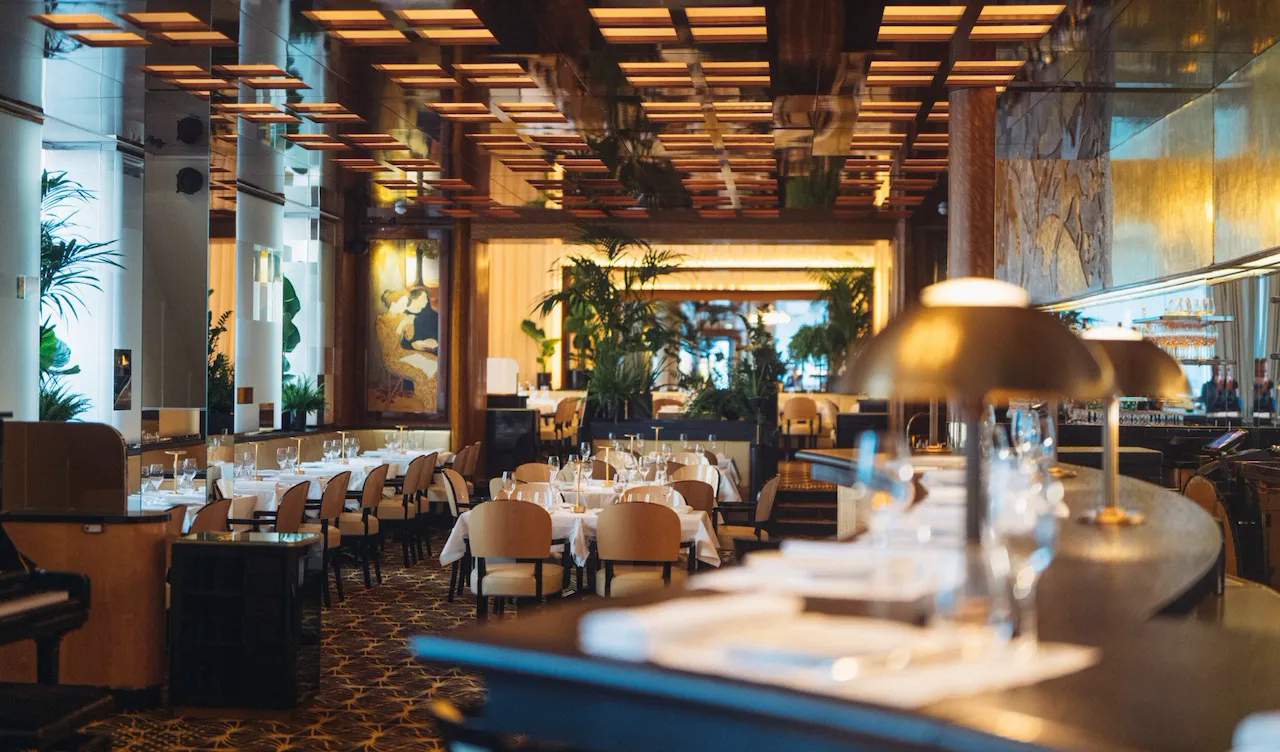 Intérieur élégant avec une ambiance chaleureuse et accueillante. Les tables sont dressées avec des nappes blanches impeccables, entourées de chaises beiges. Des plantes vertes et un éclairage tamisé renforcent l'atmosphère cosy.