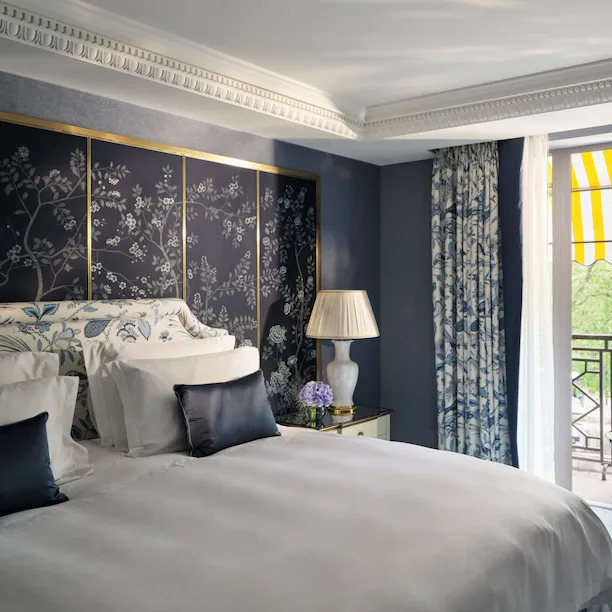 Bedroom in The Hyde Park Suite at The Dorchester, featuring blue floral décor, a king-sized bed and a private balcony with a yellow-striped awning.