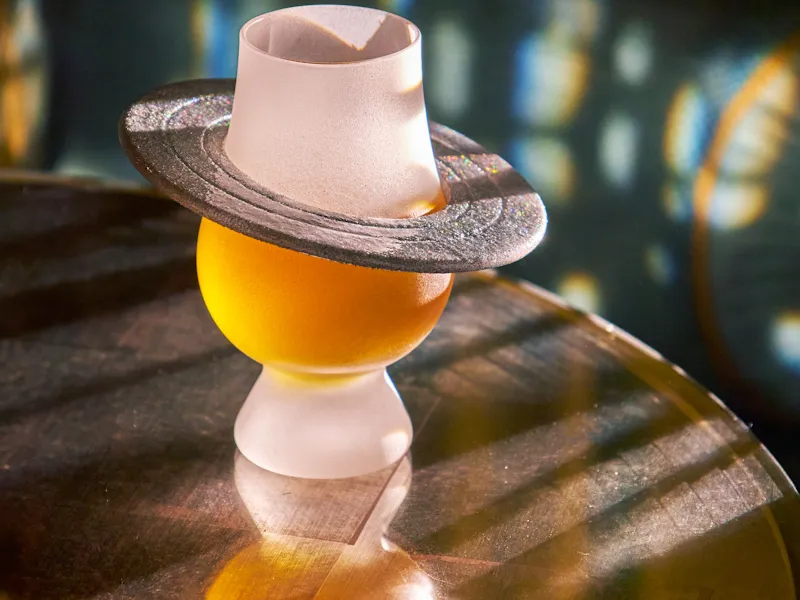 A cocktail from The Vesper Bar’s Planetarium menu, presented in a sculptural glass topped with a hat-shaped garnish on a reflective table.