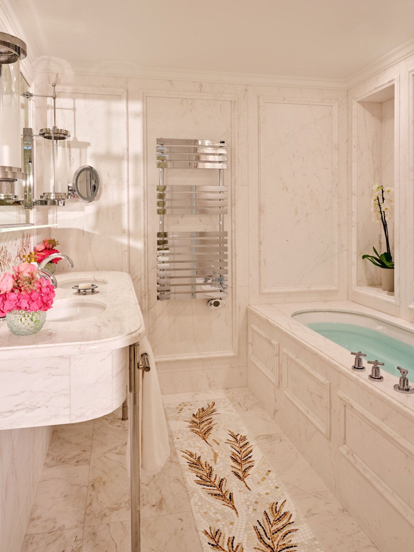 Banheiro elegante com superfícies em mármore, com pia branca decorada com flores, toalheiro cromado e banheira com decoração de orquídeas, exalando luxo.