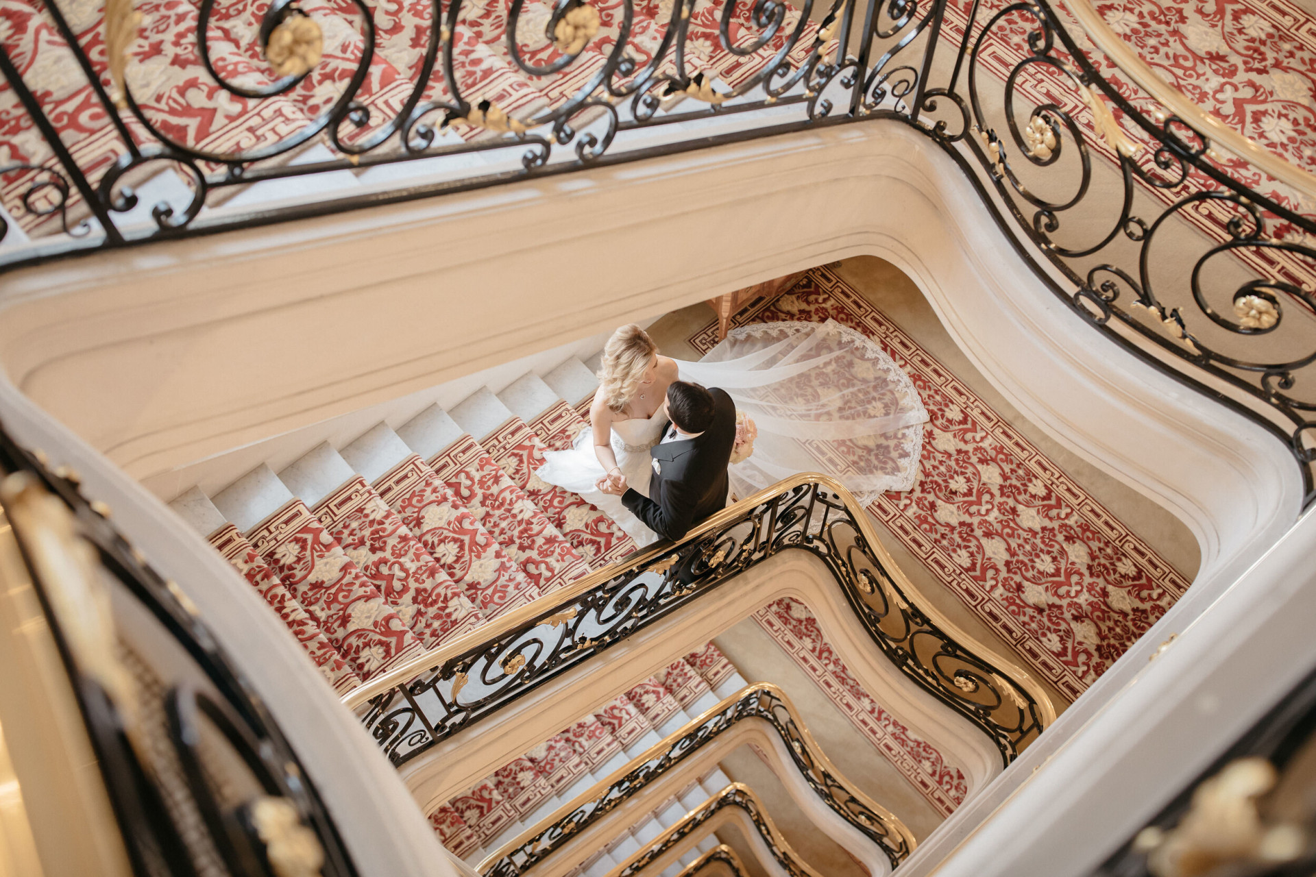 Les mariés s'embrassent sur un escalier somptueux recouvert de moquette et orné d'une rampe noire et dorée aux motifs complexes, créant une atmosphère élégante et romantique.