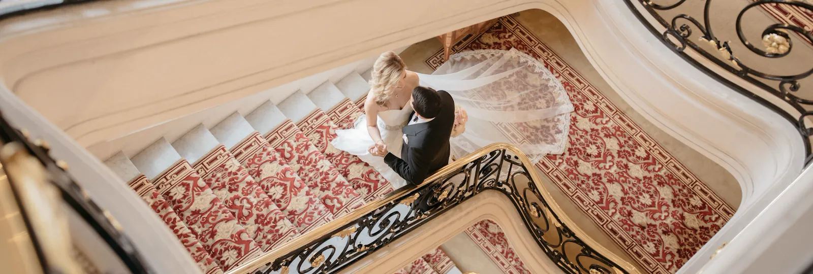 Les mariés s'embrassent sur un escalier somptueux recouvert de moquette et orné d'une rampe noire et dorée aux motifs complexes, créant une atmosphère élégante et romantique.