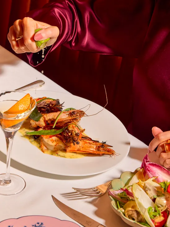 Guest squeezing fresh lime over a prawn dish at The Grill at The Dorchester, with a Martini, side salad and Ginori tableware visible.
