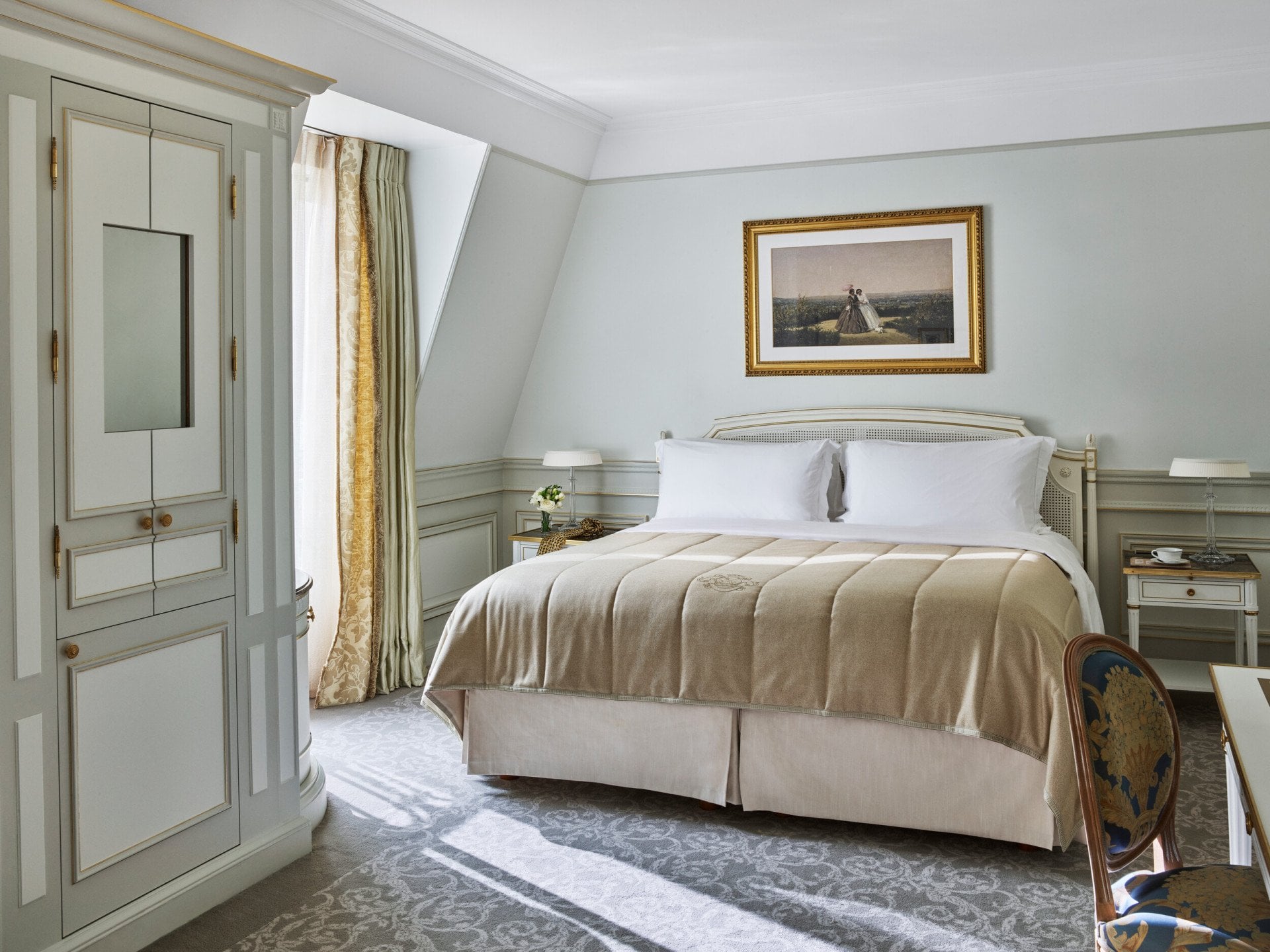 Quarto de hotel luxuoso com cama king-size bem arrumada com lençóis bege, paredes azuis claras elegantes, uma obra de arte emoldurada acima e iluminação suave.