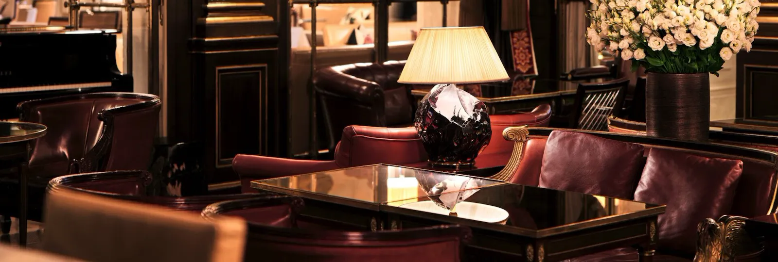 In this picture we can see three dark brown tables and five armchairs with gold details. Next to the armchairs, on a piece of furniture, is a lighted lamp with a white shade. This is reflected in one of the wooden tables. Behind one of the armchairs is a bouquet of white roses in a vase. In the background, wall lamps with white shades are lit. We can see the dark woodwork with gold detailing similar to that of the armchairs. We see a dark red curtain with gold curtain tiebacks. On the right, we see a piece of black piano. At the far end of the room we can make out the large antique windows with gold detailing overlooking another room at Le Meurice, Paris.